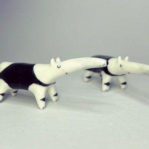 May include: Two small ceramic anteater figurines featuring a glossy white finish with black patches on the torso and legs. Each anteater has a long snout, small ears, and painted facial features including closed eyes and tiny pink cheeks.
