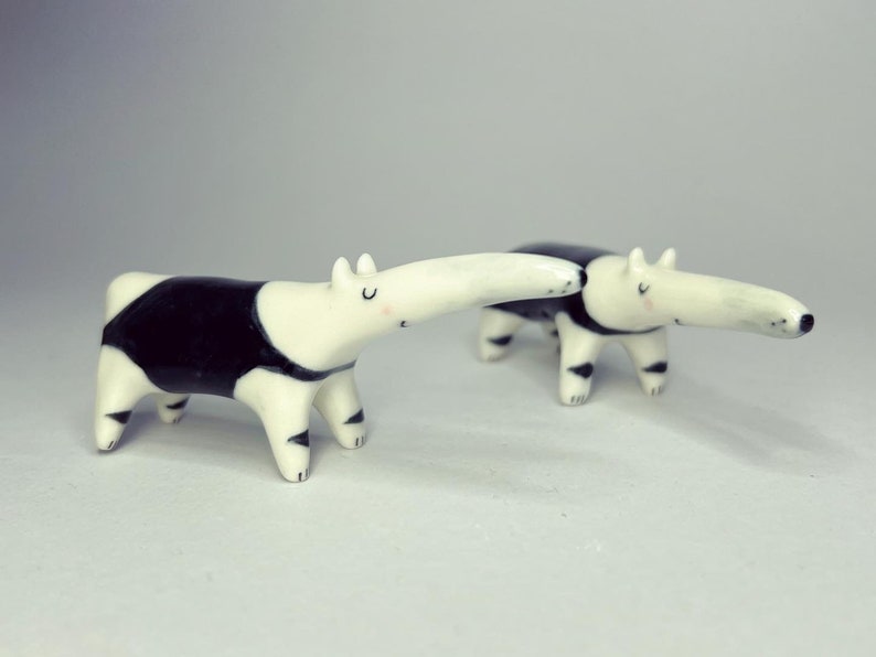 May include: Two small ceramic anteater figurines featuring a glossy white finish with black patches on the torso and legs. Each anteater has a long snout, small ears, and painted facial features including closed eyes and tiny pink cheeks.