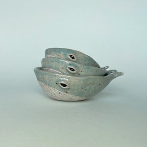 May include: Three ceramic whale-shaped bowls stacked on top of each other. The bowls are a speckled light blue with white undersides and black eye details. The bowls have a small fin-like handle.