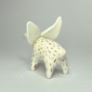 May include: A ceramic figurine of an elephant with large, upright ears. The elephant is white with brown spots and has a smooth, glossy finish. The figurine is standing on four short legs, and the ears are a lighter shade of white.