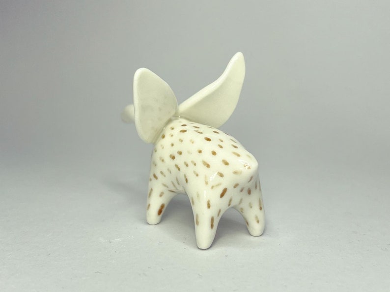 May include: A ceramic figurine of an elephant with large, upright ears. The elephant is white with brown spots and has a smooth, glossy finish. The figurine is standing on four short legs, and the ears are a lighter shade of white.
