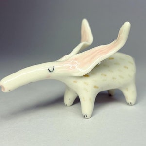 May include: A whimsical ceramic figurine of a creature with a long snout and large, upward-curving ears. The figurine is primarily off-white with pink accents on the ears and small, tan spots on the body. It has four short legs and a small, upturned tail.
