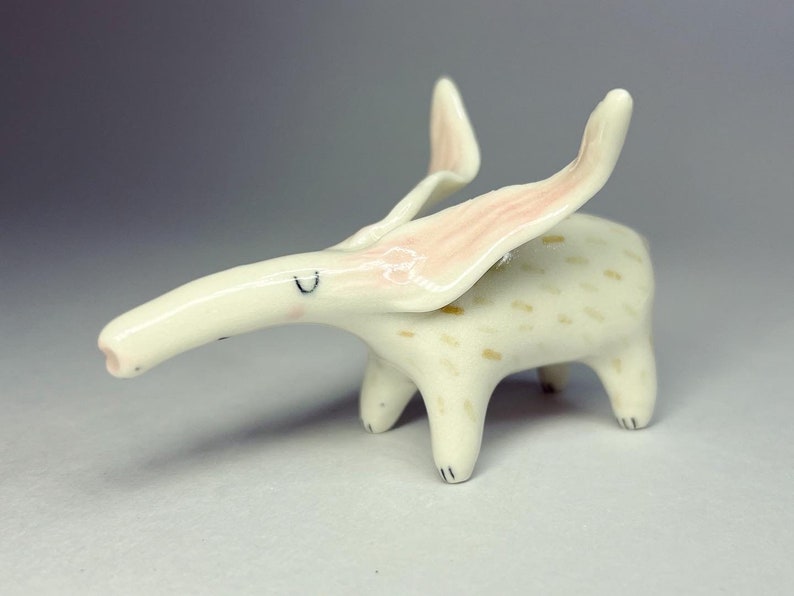 May include: A whimsical ceramic figurine of a creature with a long snout and large, upward-curving ears. The figurine is primarily off-white with pink accents on the ears and small, tan spots on the body. It has four short legs and a small, upturned tail.