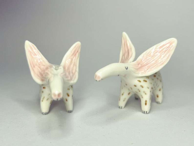 May include: Two ceramic animal figurines. The figurines are white with brown spots and large, pink-lined ears. They have long snouts and small, black eyes. The figurines are standing on four legs.