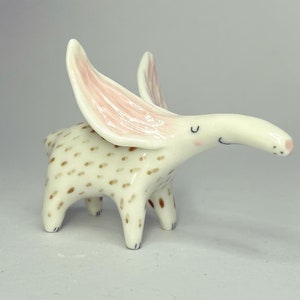 May include: A whimsical ceramic figurine of a creature with large, pink-lined ears. The off-white body is speckled with brown spots. The creature has a long snout and small, black eyes. The figurine is standing on four short legs.
