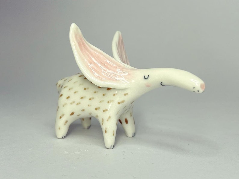 May include: A whimsical ceramic figurine of a creature with large, pink-lined ears. The off-white body is speckled with brown spots. The creature has a long snout and small, black eyes. The figurine is standing on four short legs.