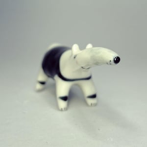 May include: A small ceramic figurine of a tapir with a smooth, glossy finish. The animal is primarily white with a distinct black patch covering its midsection and back, and black markings on its legs. It has a long snout and a small black nose.