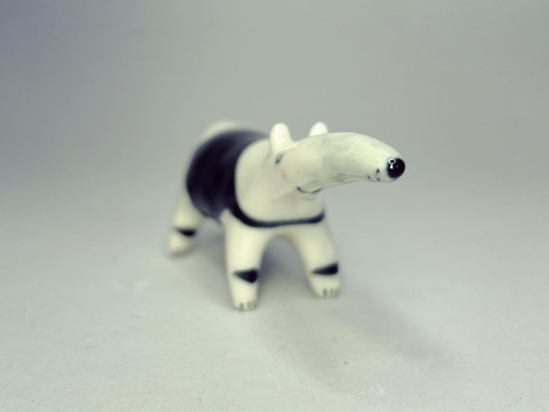 May include: A small ceramic figurine of a tapir with a smooth, glossy finish. The animal is primarily white with a distinct black patch covering its midsection and back, and black markings on its legs. It has a long snout and a small black nose.