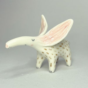 May include: A whimsical ceramic figurine of a creature with long ears and a curved snout. The figurine is primarily white with brown spots on its body and pink accents inside the ears. It has four legs and a small tail.