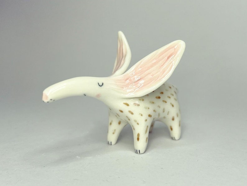 May include: A whimsical ceramic figurine of a creature with long ears and a curved snout. The figurine is primarily white with brown spots on its body and pink accents inside the ears. It has four legs and a small tail.