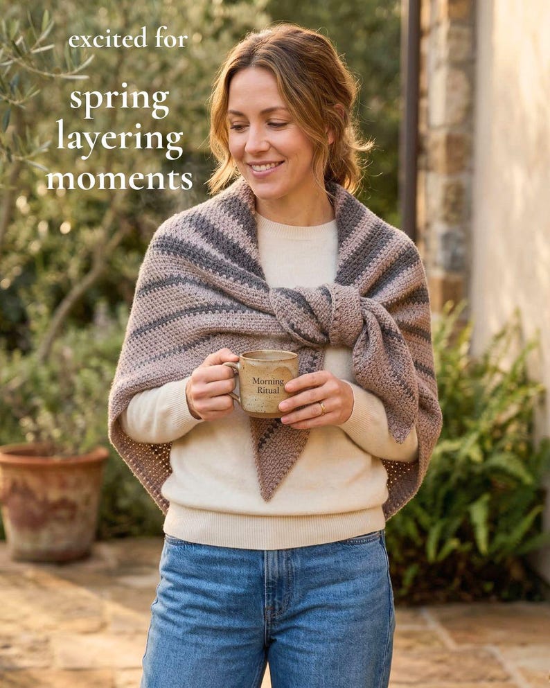 May include: A woman wearing a beige sweater and blue jeans, with a striped shawl in shades of gray and beige. She holds a ceramic mug with the words "Morning Ritual" printed on it. The text "excited for spring layering moments" is in the upper left corner.