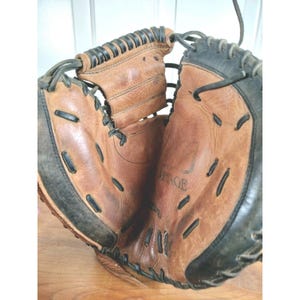 May include: A tan and black leather baseball catcher's mitt featuring thick black lacing along the edges and webbed sections. The interior palm area displays a visible embossed logo with the word VINTAGE centered on the worn leather surface.