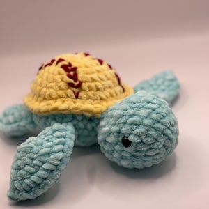 May include: A handmade crochet turtle in shades of light blue and yellow. The turtle's shell is yellow with maroon stitching, and the body and flippers are light blue. The turtle has a black eye.
