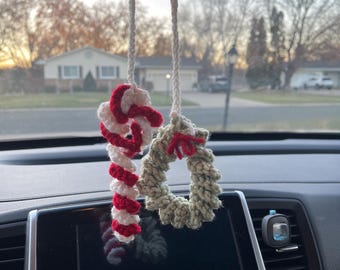 May include: Two crocheted Christmas decorations hanging from a car's rearview mirror. One is a red and white candy cane, the other a green wreath with a red bow. The ornaments are suspended by white cords.