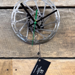 May include: A unique wall clock made from a bicycle disc brake rotor. The clock features black hands, a green second hand, and a silver-colored metal finish. The clock is mounted on a wooden background. A black tag hangs below the clock.