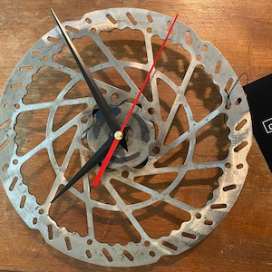 May include: A unique clock made from a silver-colored bicycle disc brake rotor. The clock features black hour and minute hands, a red second hand, and a gold-colored center. A black tag is attached to the clock.