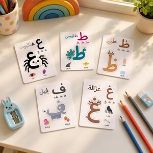 Puede incluir: Cinco tarjetas educativas rectangulares blancas con letras árabes, cada una acompañada de una ilustración de un animal u objeto y texto. Las tarjetas muestran una araña, un pavo real, una gacela, un elefante y una criatura similar a un ciervo.