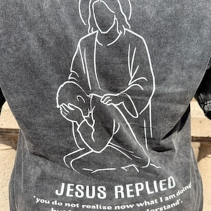 May include: Dark gray t-shirt featuring a white line drawing of Jesus comforting another figure. The text "JESUS REPLIED" and a quote are printed below the illustration. The shirt has a vintage wash.