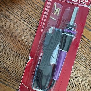 Puede incluir: Un kit de pirograbado en madera en un paquete rojo. El bolígrafo tiene un mango morado y una punta plateada, con un cable de alimentación negro. Incluye varias puntas intercambiables.