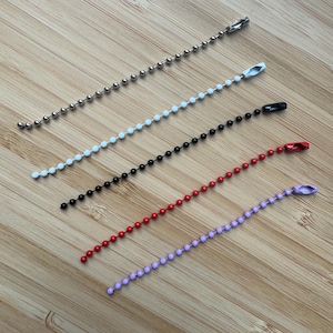 May include: Five ball chains in silver, white, black, red, and light purple. Each chain features small spherical beads connected in a line and is finished with a matching metal connector clasp at one end for securing the ends together.
