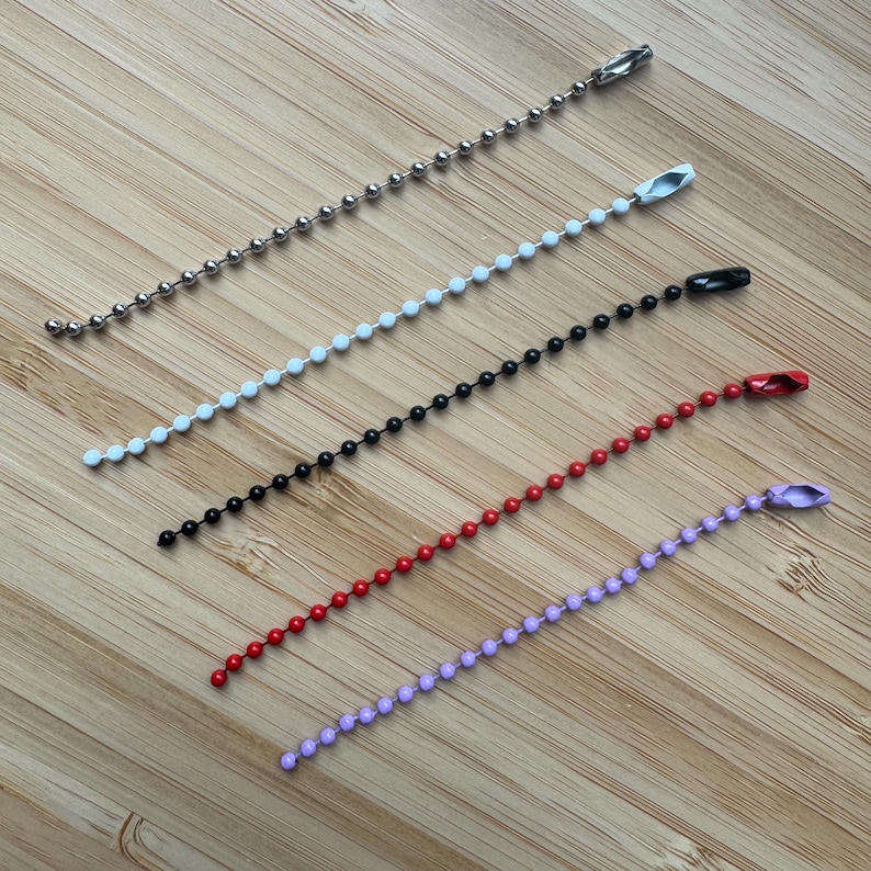 May include: Five ball chains in silver, white, black, red, and light purple. Each chain features small spherical beads connected in a line and is finished with a matching metal connector clasp at one end for securing the ends together.