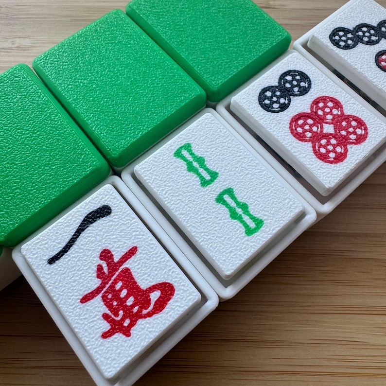 May include: A collection of Mahjong tiles featuring white faces with engraved symbols in red, green, and black ink. The tiles have bright green textured backs and white plastic frames, displaying characters and circular patterns on their front surfaces.
