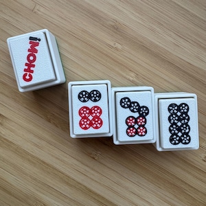 May include: Four rectangular white mahjong tiles with raised edges. One tile features the red text CHOW! vertically. The other three tiles display circular patterns in black and red ink, representing numerical values with dotted designs on their faces.