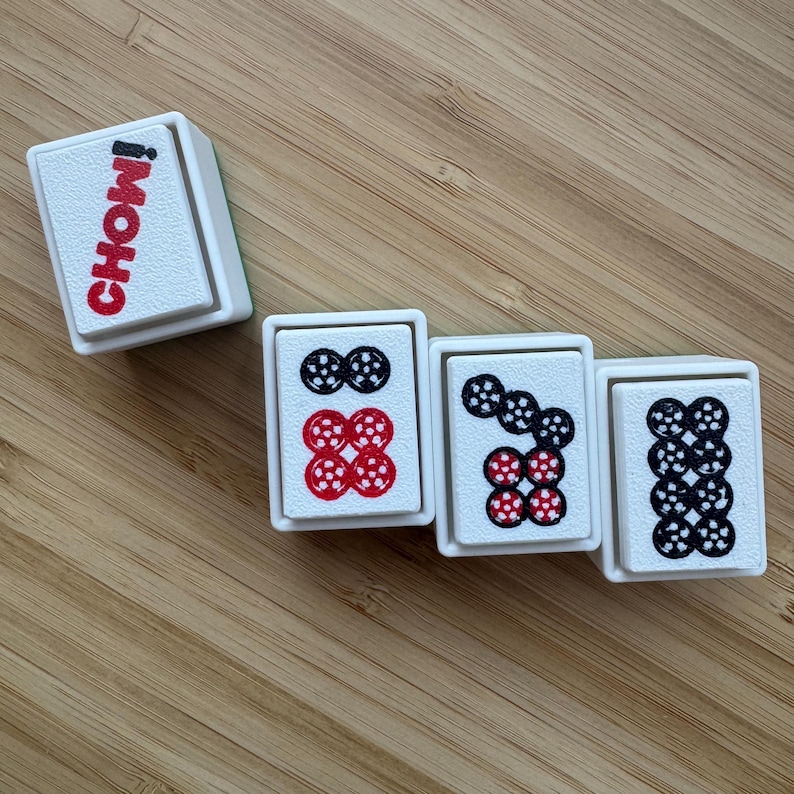 May include: Four rectangular white mahjong tiles with raised edges. One tile features the red text CHOW! vertically. The other three tiles display circular patterns in black and red ink, representing numerical values with dotted designs on their faces.