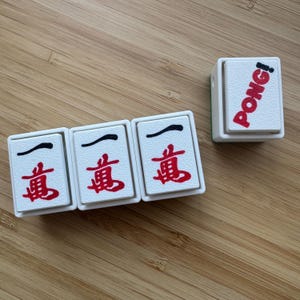 May include: Three white rectangular mahjong tiles featuring the red and black Chinese character for ten thousand, arranged in a row, alongside a single white tile standing upright with the word PONG! printed in red and black lettering.