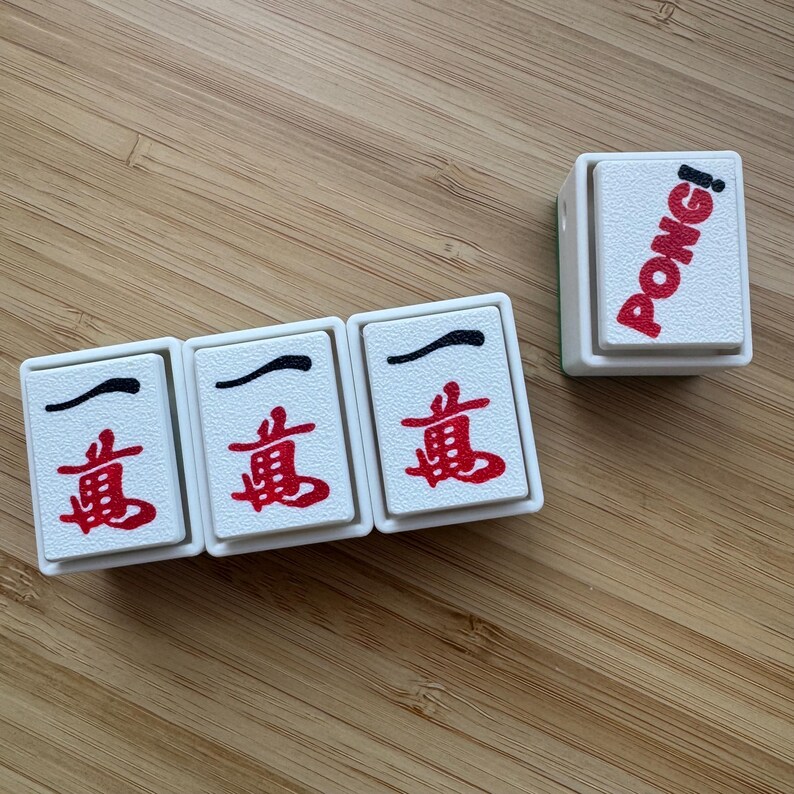 May include: Three white rectangular mahjong tiles featuring the red and black Chinese character for ten thousand, arranged in a row, alongside a single white tile standing upright with the word PONG! printed in red and black lettering.