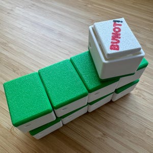 May include: A set of rectangular blocks with white bases and textured green tops, arranged in a staggered formation. The top block features a white lid with the red text BUNOT! printed on it. Each block has a small circular hole on the side.