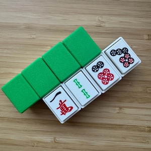 May include: A set of rectangular mahjong tiles arranged in a diagonal row. Three tiles have solid green backs, while three white-faced tiles display red and black ink markings, including a red character, green bamboo symbols, and circular patterns.