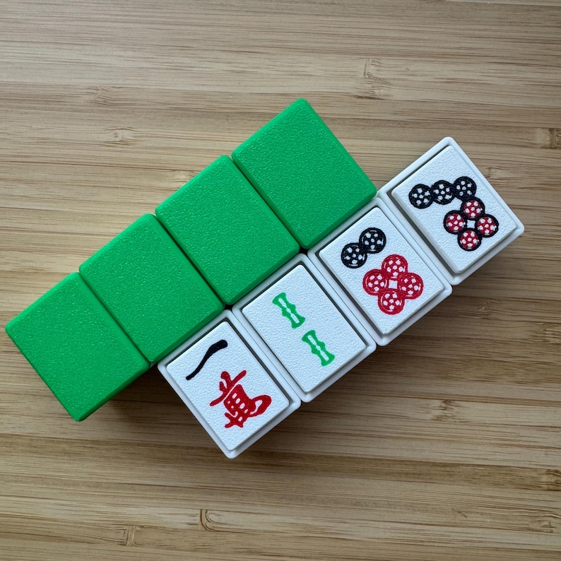 May include: A set of rectangular mahjong tiles arranged in a diagonal row. Three tiles have solid green backs, while three white-faced tiles display red and black ink markings, including a red character, green bamboo symbols, and circular patterns.