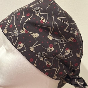 May include: A dark gray scrub cap featuring a repeating pattern of off-white skeletons with red flowers tucked behind their ears. The fabric head covering has a wide band and a tie-back closure at the nape, displaying a consistent skeletal print.