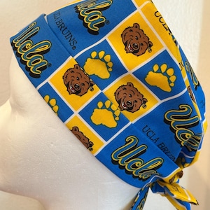 May include: Blue and gold patterned scrub cap featuring the UCLA Bruins logo, bear faces, and paw prints. The cap is tied at the back and is made of a soft, likely cotton, fabric. The text "UCLA Bruins" is visible.