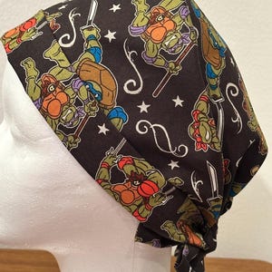 May include: A black head covering featuring a repeating pattern of the Teenage Mutant Ninja Turtles in various action poses. The design includes white stars and swirls. The head covering is tied at the back.