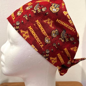 May include: A red and gold patterned head covering featuring the word "Minnesota" and a cartoon gopher mascot. The head covering is tied at the back and is displayed on a white mannequin head.