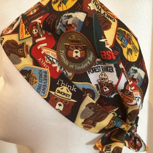 May include: A black and yellow patterned scrub cap featuring Smokey Bear and various forest-themed patches. The cap includes text such as "Prevent Forest Fires" and "Only You". The cap is tied at the back.