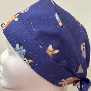 May include: A dark blue scrub cap featuring a repeating pattern of cartoon dogs in shades of orange, blue, and pink against a background of small white dots. The fabric head covering has a wide band and is tied at the back with fabric ends.