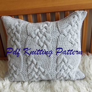 May include: A gray knitted pillow with a cable knit pattern. The pillow is sitting on a white fluffy surface.