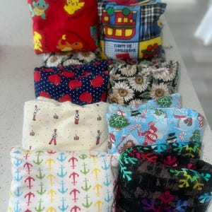 Puede incluir: Una colección de bolsas de tela coloridas con varios patrones. Los diseños incluyen personajes de dibujos animados, manzanas, girasoles, soldados de juguete, escenas navideñas y anclas. Las bolsas están dispuestas en filas, mostrando las diversas estampados de tela.