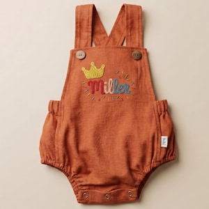 May include: Rust-colored baby romper with suspender straps and button closures. The romper features an embroidered gold crown and the name "Miller" in colorful thread. A small white tag is sewn on the side.