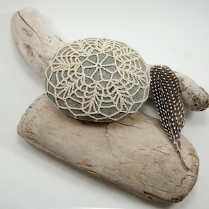 May include: A round, gray stone covered in a delicate, off-white crocheted lace pattern. The stone is resting on two pieces of driftwood, one of which has a brown and white speckled feather resting on top.