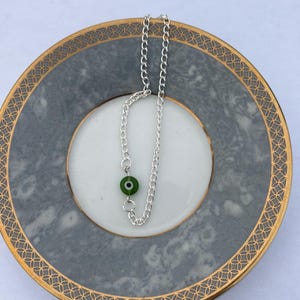May include: A silver chain necklace with a green evil eye pendant. The necklace is displayed on a gray and white plate with a gold rim. The evil eye pendant is a dark green color with a black center.