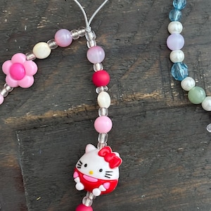 May include: A beaded phone charm featuring a pink flower, pink and white beads, and a Hello Kitty charm. The charm has a clear string for attaching to a phone or other device. The beads are various sizes and shapes.