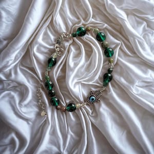 May include: A beaded bracelet featuring dark green and light green beads, interspersed with smaller, lighter-colored beads. A silver-toned Hamsa hand charm with a blue eye detail is attached. The bracelet is displayed on a white, silky fabric.