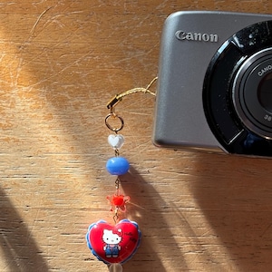 May include: A silver Canon digital camera with a decorative charm. The charm features a red heart-shaped pendant with a Hello Kitty design, blue and white beads, and a gold-colored chain.
