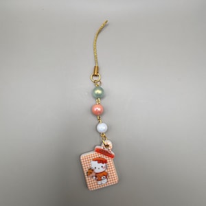 May include: A gold-colored charm with a string of pastel-colored beads and a small, square charm featuring a Hello Kitty design. The charm has a gold-colored string and a small tag that says "HERMES".