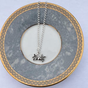 May include: A silver-colored necklace featuring a bird pendant holding a sprig of leaves. The necklace is displayed on a gray and white plate with a gold rim. The chain is delicate and hangs from the pendant.