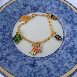 May include: Gold-toned charm bracelet featuring colorful enamel charms. Charms include a green leaf, a blue leaf, a yellow umbrella, and a charm of two figures in orange coats. The bracelet is displayed on a blue and white plate.